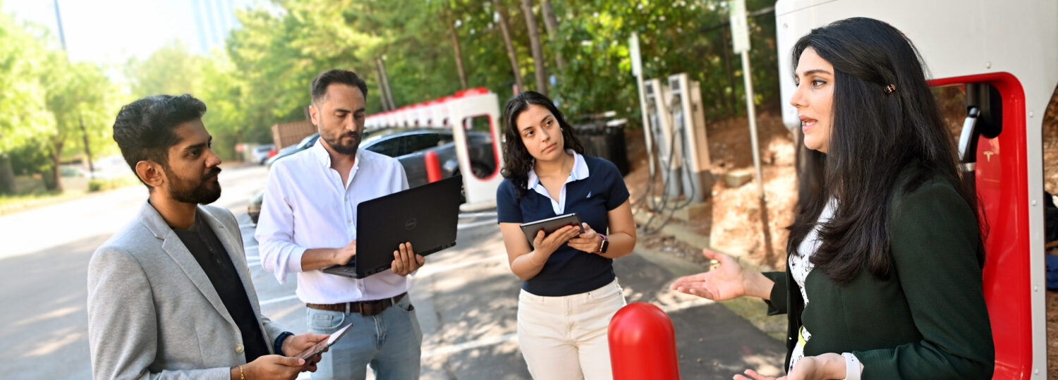 Leila Hajibabai talking with three students while standing in front of an electric car charging station.