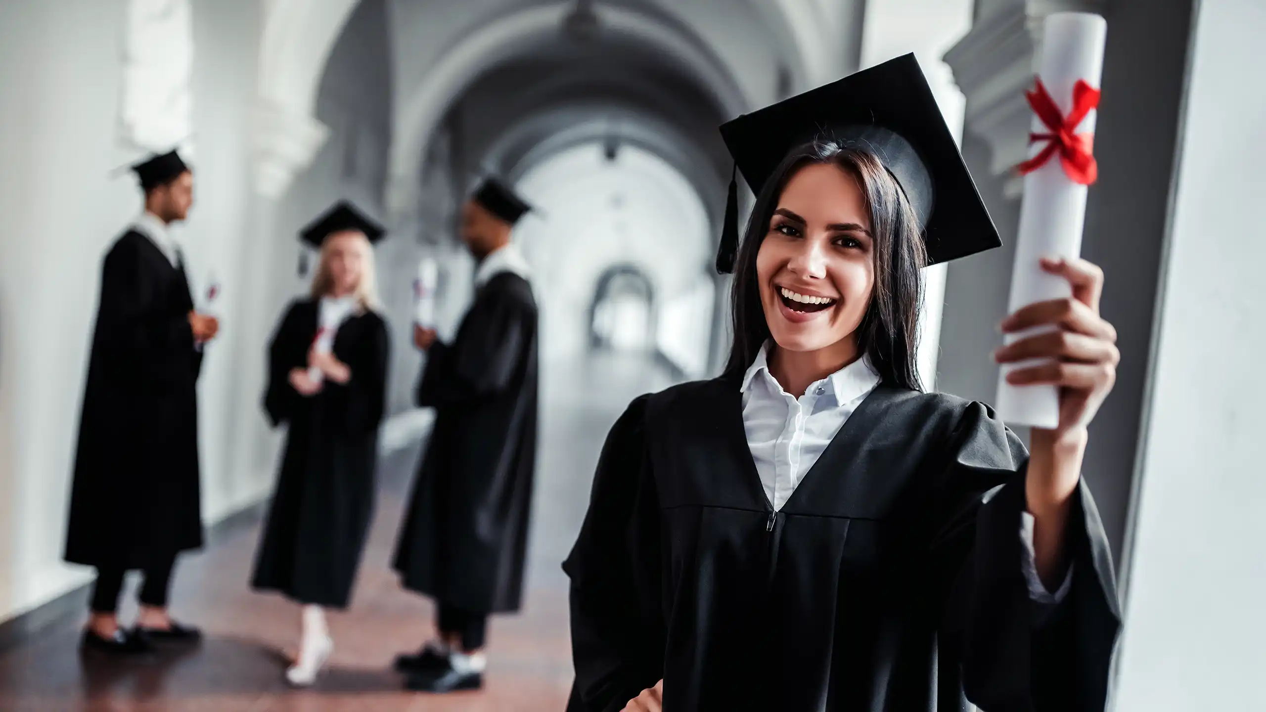 A female student standing in the hallway of a large building holding up her degree.