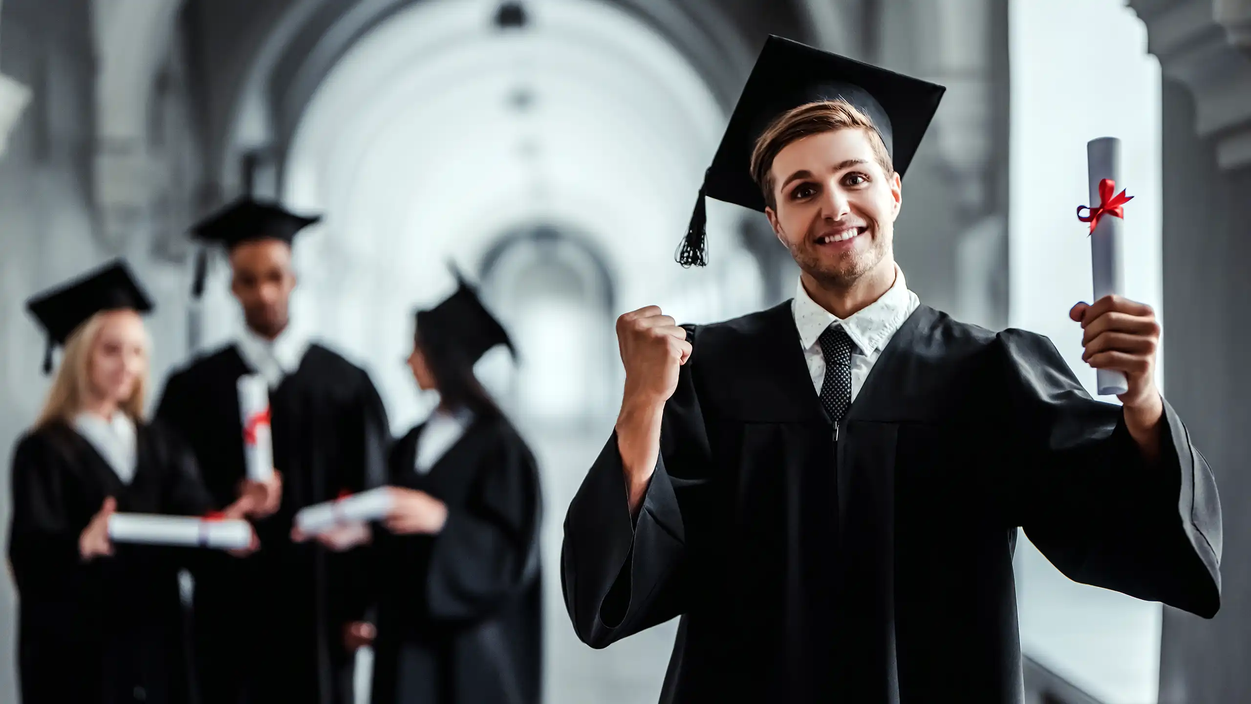 A male student is standing in the hallway of a large building, holding up his degree and pumping his fist in excitement.