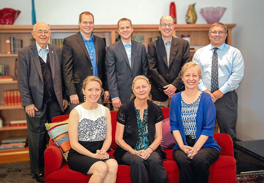 Richard Bernhard is standing behind a couch with his sons and sons-in-law. On the couch is his wife Cindy and his daughter and daughter-in-law.