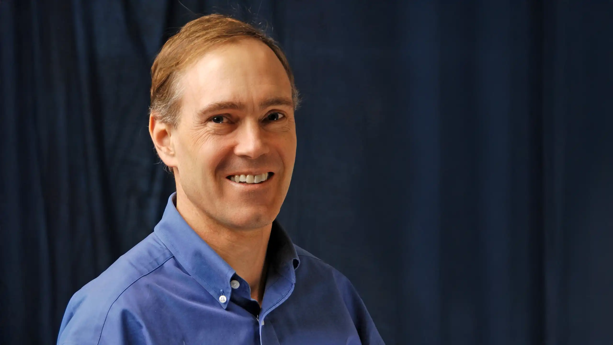 A headshot of John Lee standing in front of a dark blue curtain.