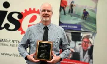 Russell "rusty" King stands in front of a giant ISE Departmental sign holding his seventh Clifton A. Anderson Outstanding Faculty Award
